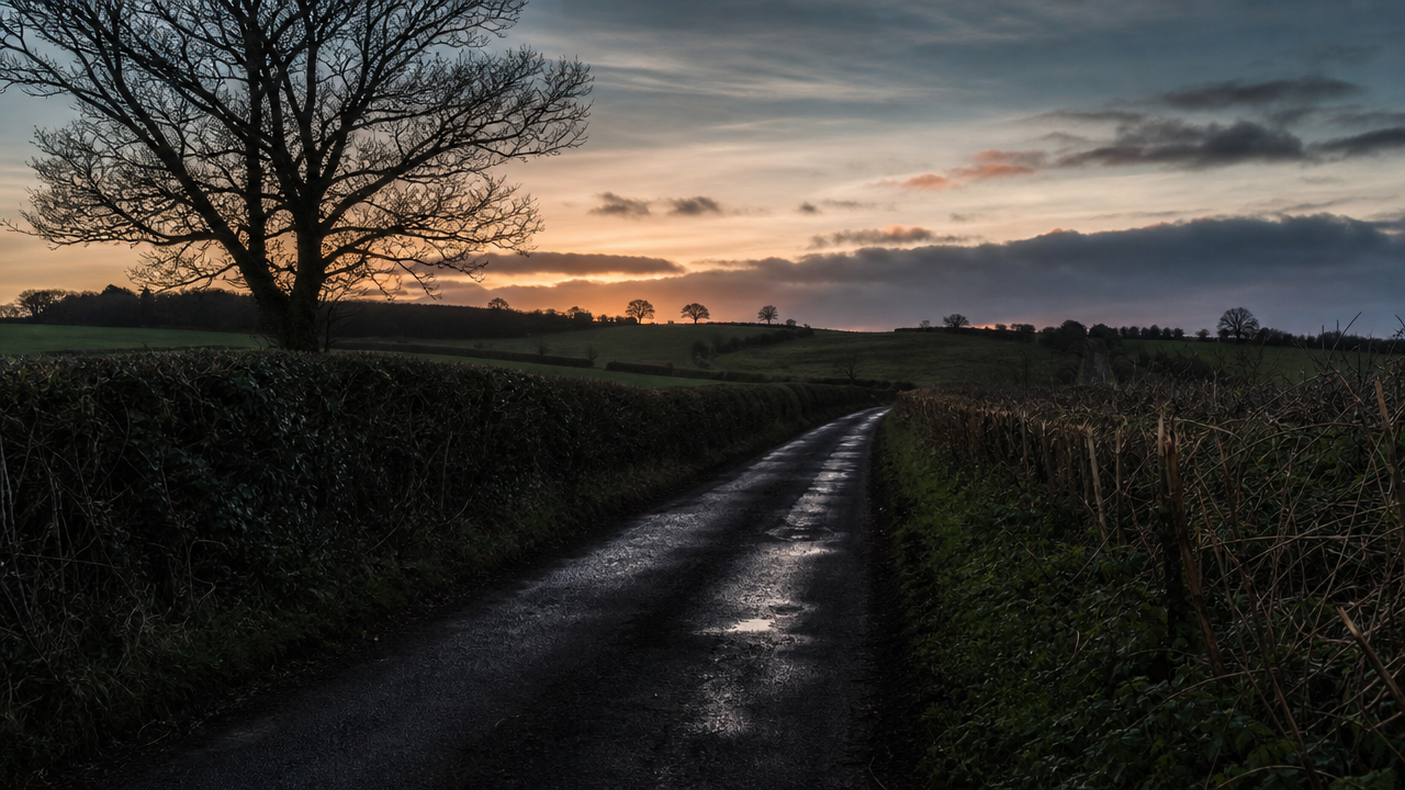 An empty dusk road with a single damp lane stretching into the distance. FACEBOOK ANGLE: The strangest part was not that it rained. It was that the witnesses said it chose a lane. FACEBOOK VISUAL MOMENT: A clean dark stripe of wet pavement running down one side of a country road while the other lane and shoulders stayed dry. FACEBOOK SHORT SUMMARY: Near Ashford, several witnesses described rain falling on only one lane of a rural road for several minutes. The later wet stripe had no obvious sprinkler, tanker, or drainage explanation.
