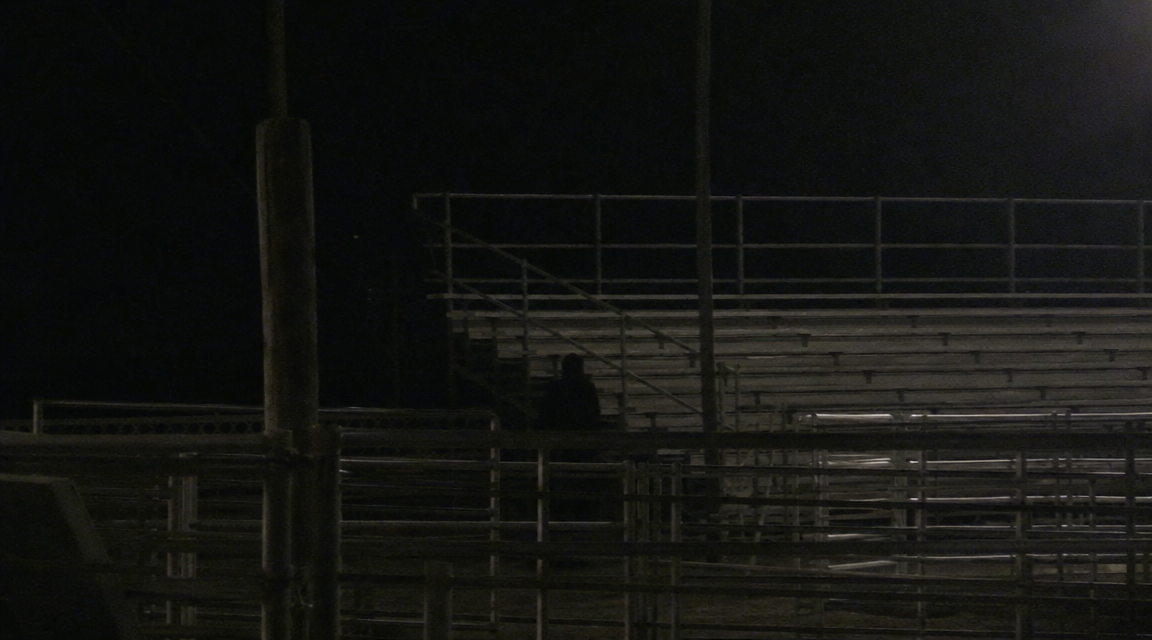 Low-light fairground image with a broad dark upright shape partly hidden behind rail shadows near empty bleachers.