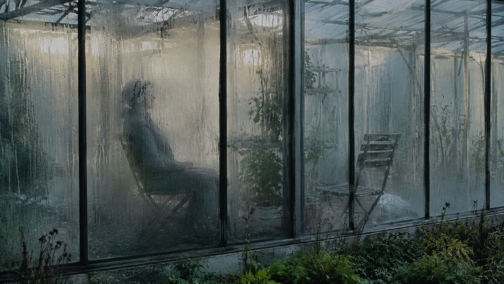 Condensation gathers in a seated outline beside an empty chair in a cold glasshouse. FACEBOOK ANGLE: The haunting at Larkmere Orchard is unsettling because it behaves like a room still being used, not a place trying to frighten anyone. FACEBOOK VISUAL MOMENT: A cold pane inside the ruined glasshouse slowly clouds over, leaving a warm palm print where no hand has touched it. FACEBOOK SHORT SUMMARY: Visitors to Larkmere Orchard’s derelict glasshouse report handprints appearing on the inside of cold panes, an old chair changing its view, and condensation shaping itself around an unseen sitter.