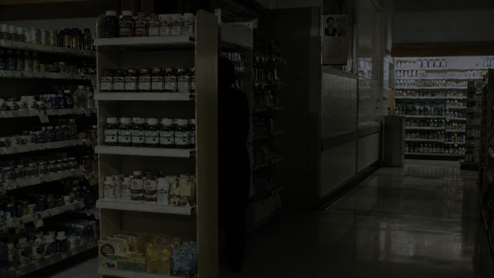 Dim pharmacy endcap with an indistinct dark figure-like shape partly hidden behind the vitamin display.