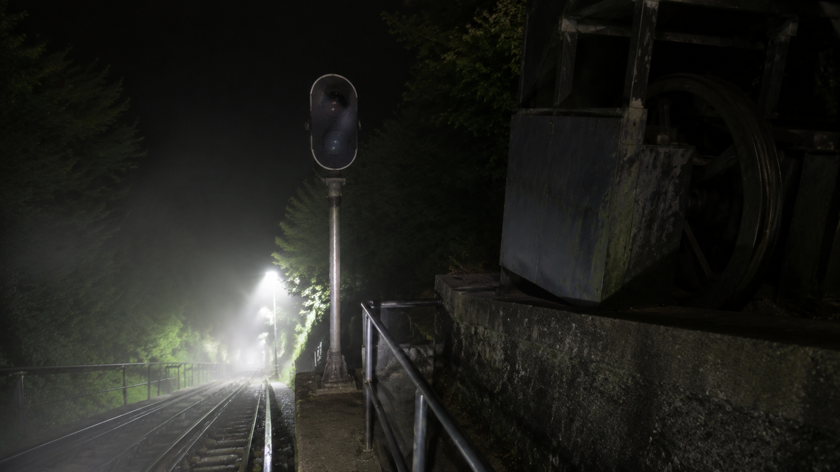 Misty nighttime funicular platform with rails, railing, and an unlit signal lamp.