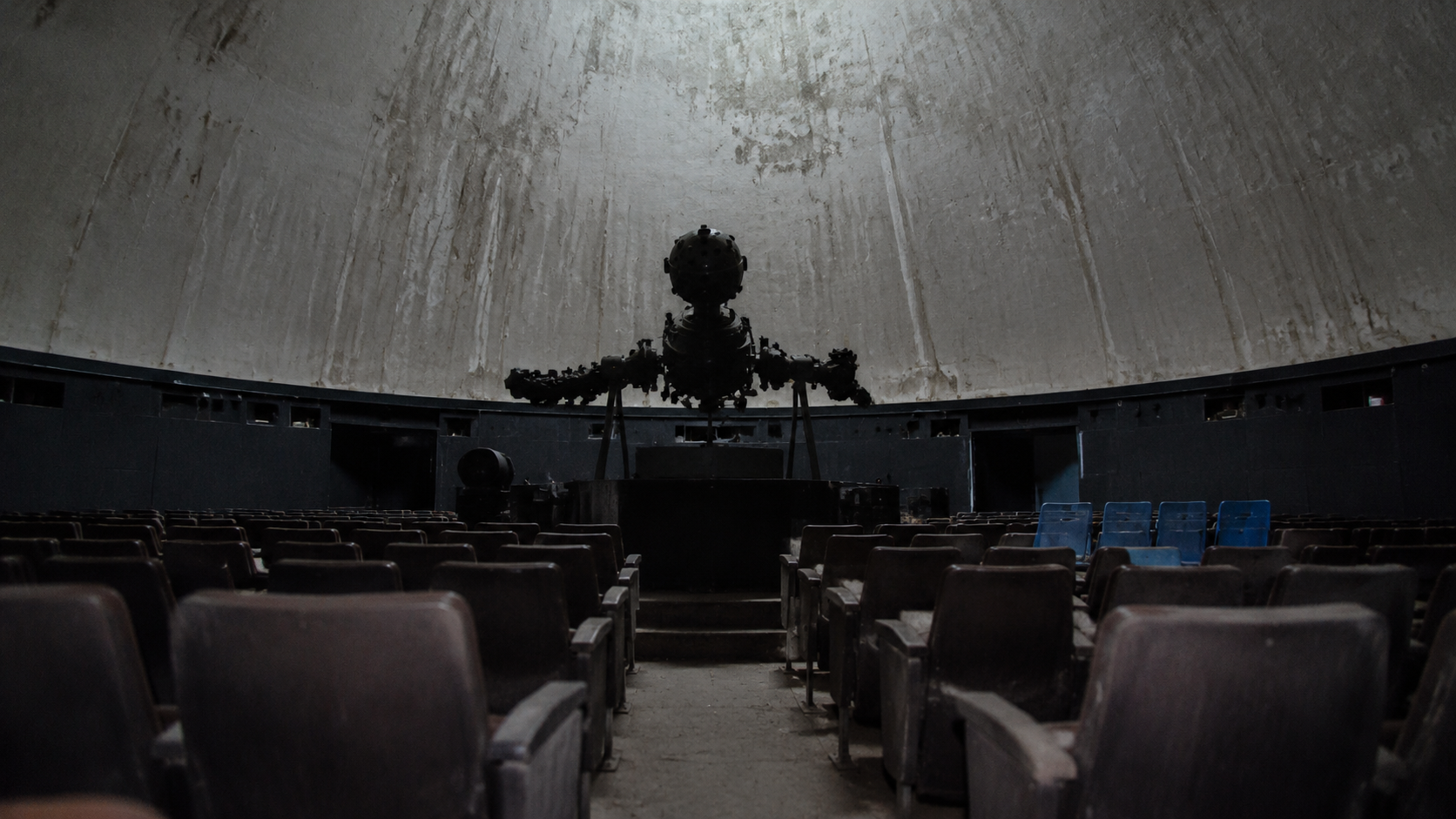 The back row of an abandoned planetarium looks toward a silent star projector under a stained dome. FACEBOOK ANGLE: A closed civic planetarium should have been uniformly dusty, but several chair backs looked recently used. FACEBOOK VISUAL MOMENT: The old star projector still faces the seats while a small cluster of clean chair backs interrupts the dust. FACEBOOK SHORT SUMMARY: Maintenance workers entering a roof-damaged planetarium found the room arranged like a show had just ended, with clean chair backs in an otherwise dusty theater.