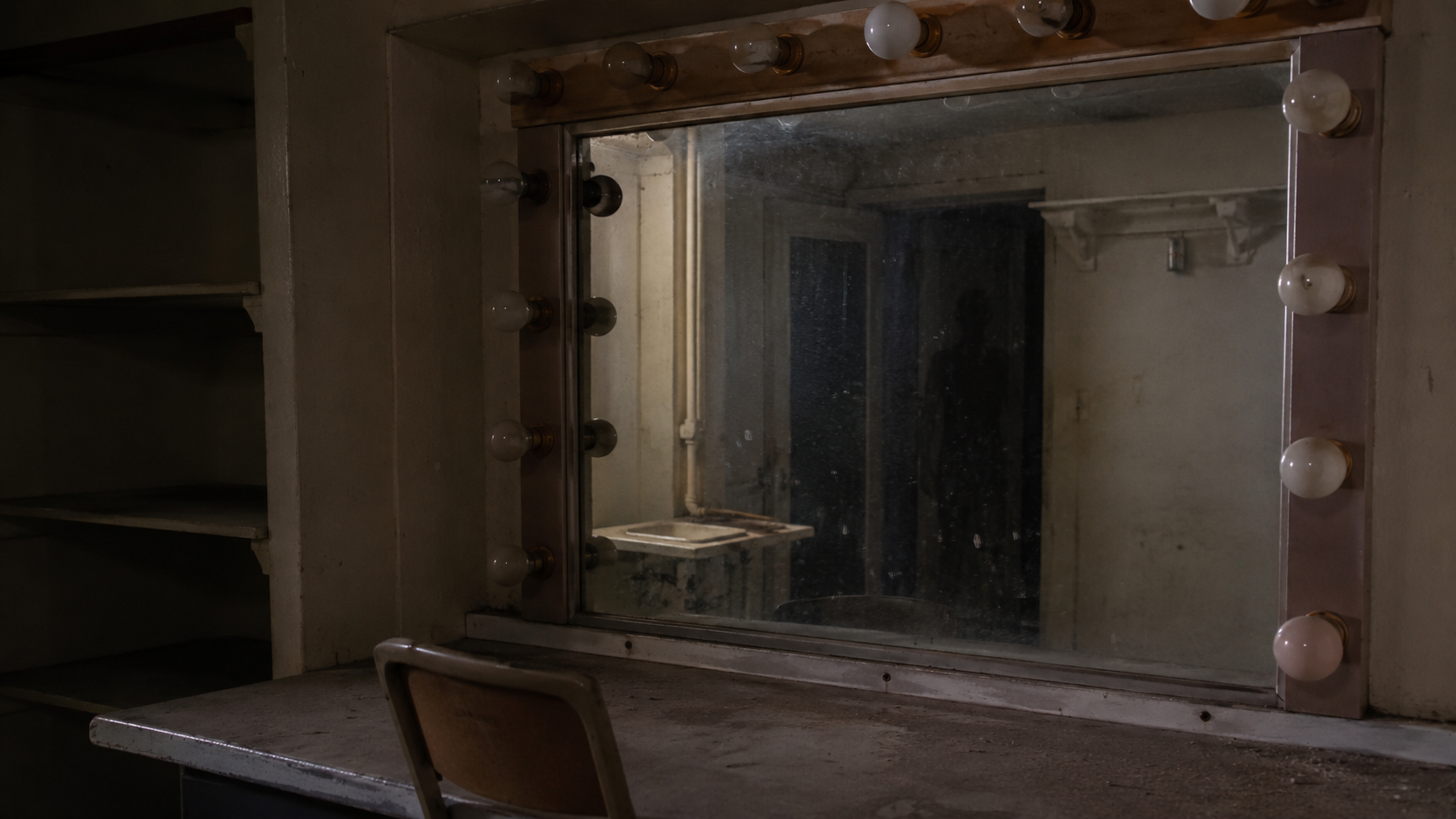 A dusty dressing room mirror with a faint dark shape near the edge of the reflection. FACEBOOK ANGLE: A locked dressing room under a closed theater has mirror bulbs that still glow after the circuit was marked dead. FACEBOOK VISUAL MOMENT: The maintenance photo where the mirror shows the room, the bulbs, and a dark human-shaped shape standing just inside the reflected edge. FACEBOOK SHORT SUMMARY: Beneath the Orpheum, a forgotten dressing room keeps drawing attention for one simple reason: it behaves as if someone is still using it.