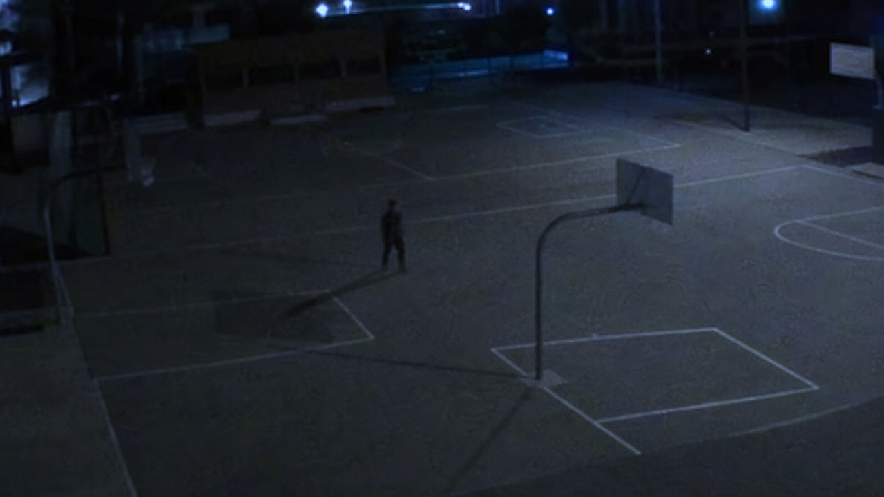 A narrow indistinct figure standing near a basketball hoop shadow in a closed schoolyard.
