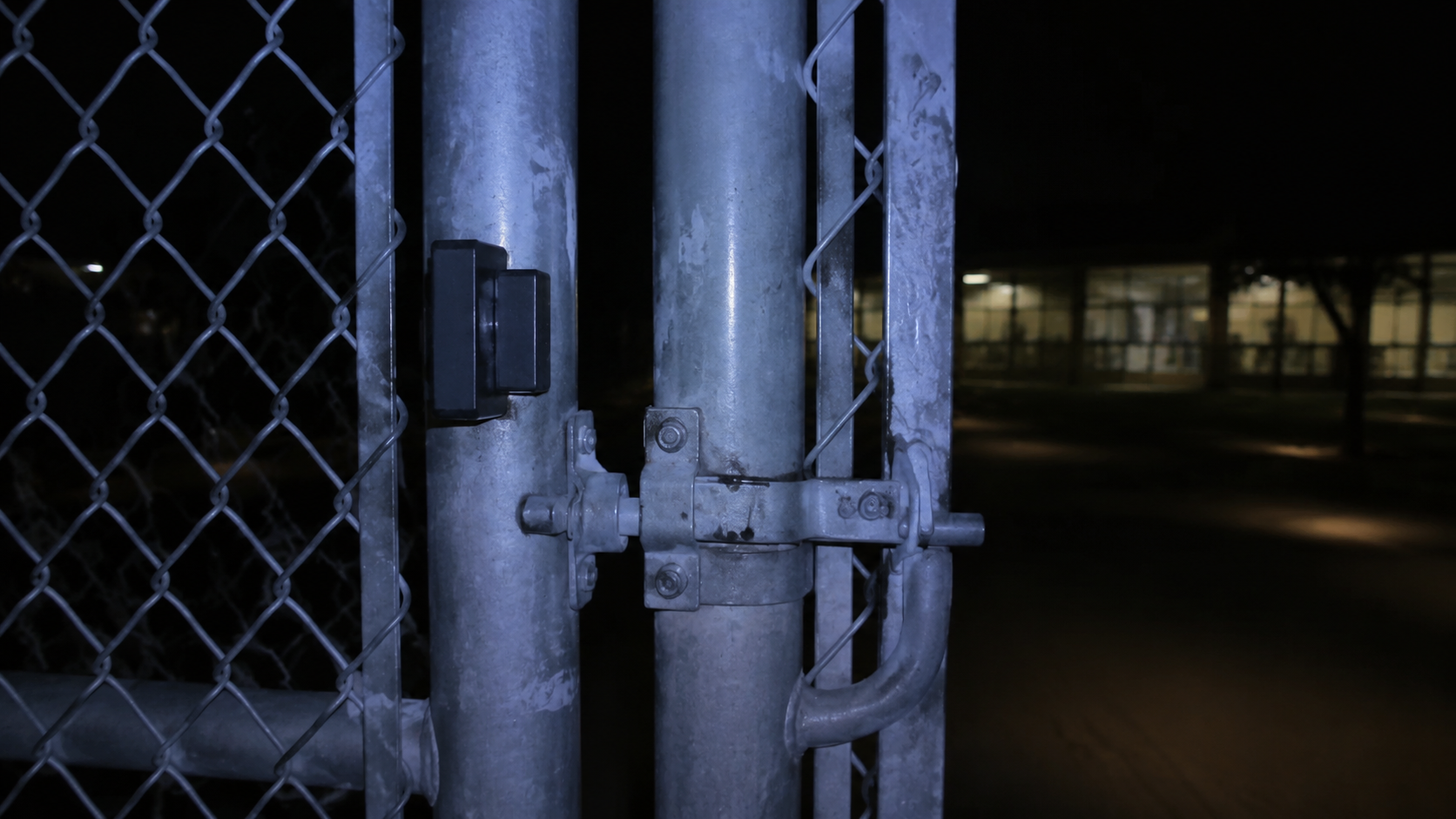 A closed schoolyard gate latch and magnetic sensor under blue-white night light.