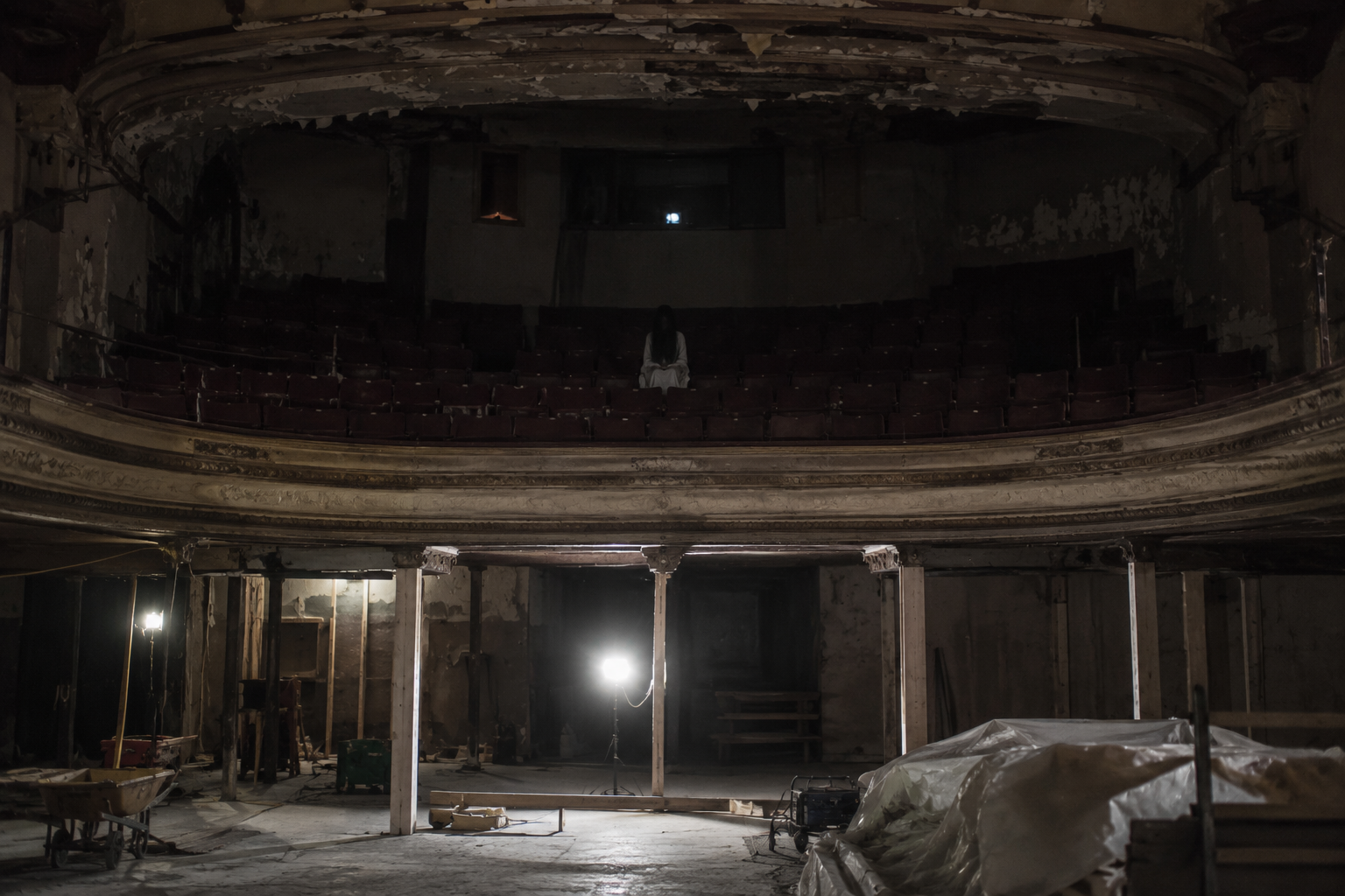 Work lights shine across an abandoned theater while a pale figure appears among the balcony seats.