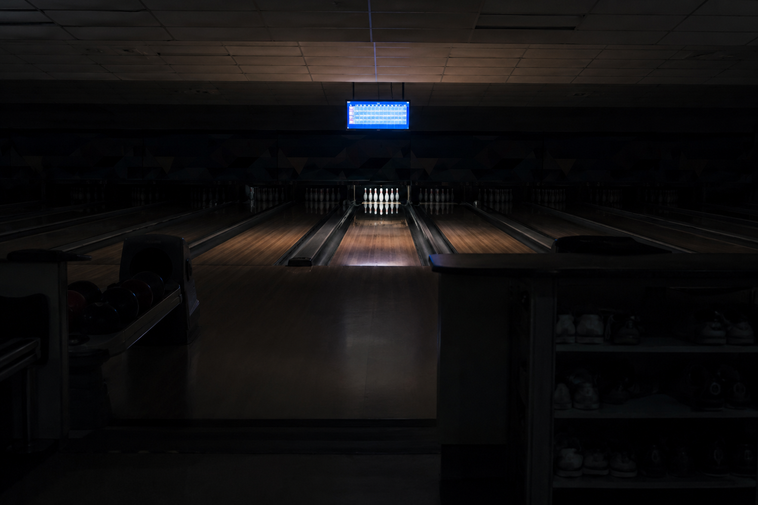 Shadowy bowling alley concourse with one distant overhead scoreboard turned on over an empty lane.