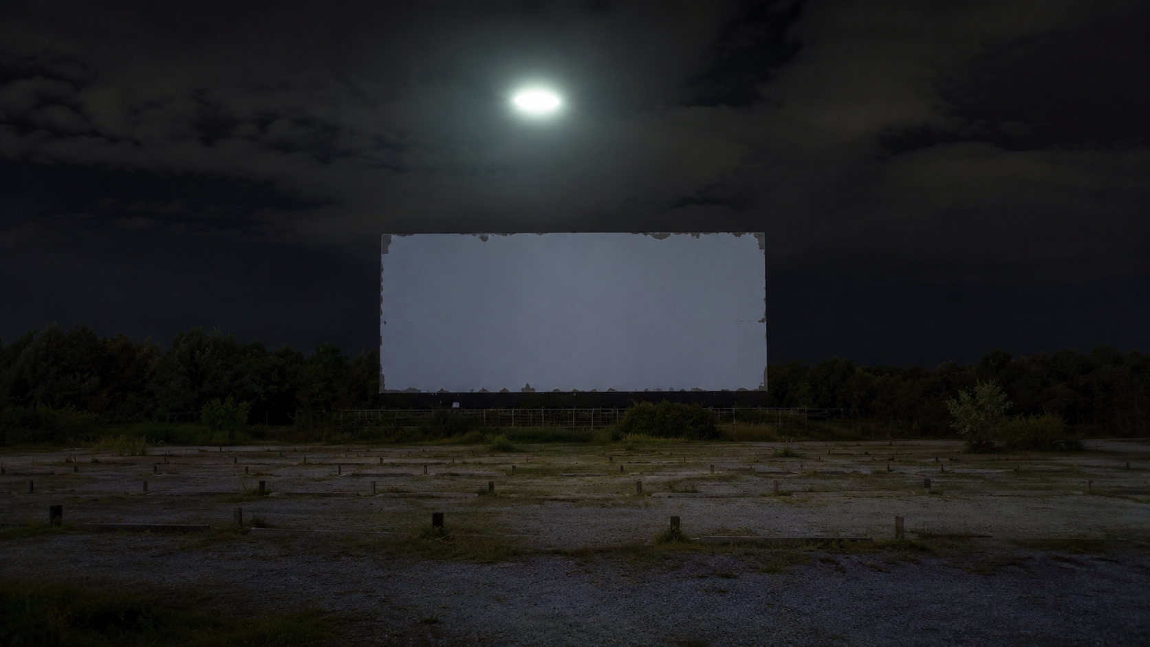 A pale oval light hovers above the blank screen at a closed rural drive-in theater after midnight.