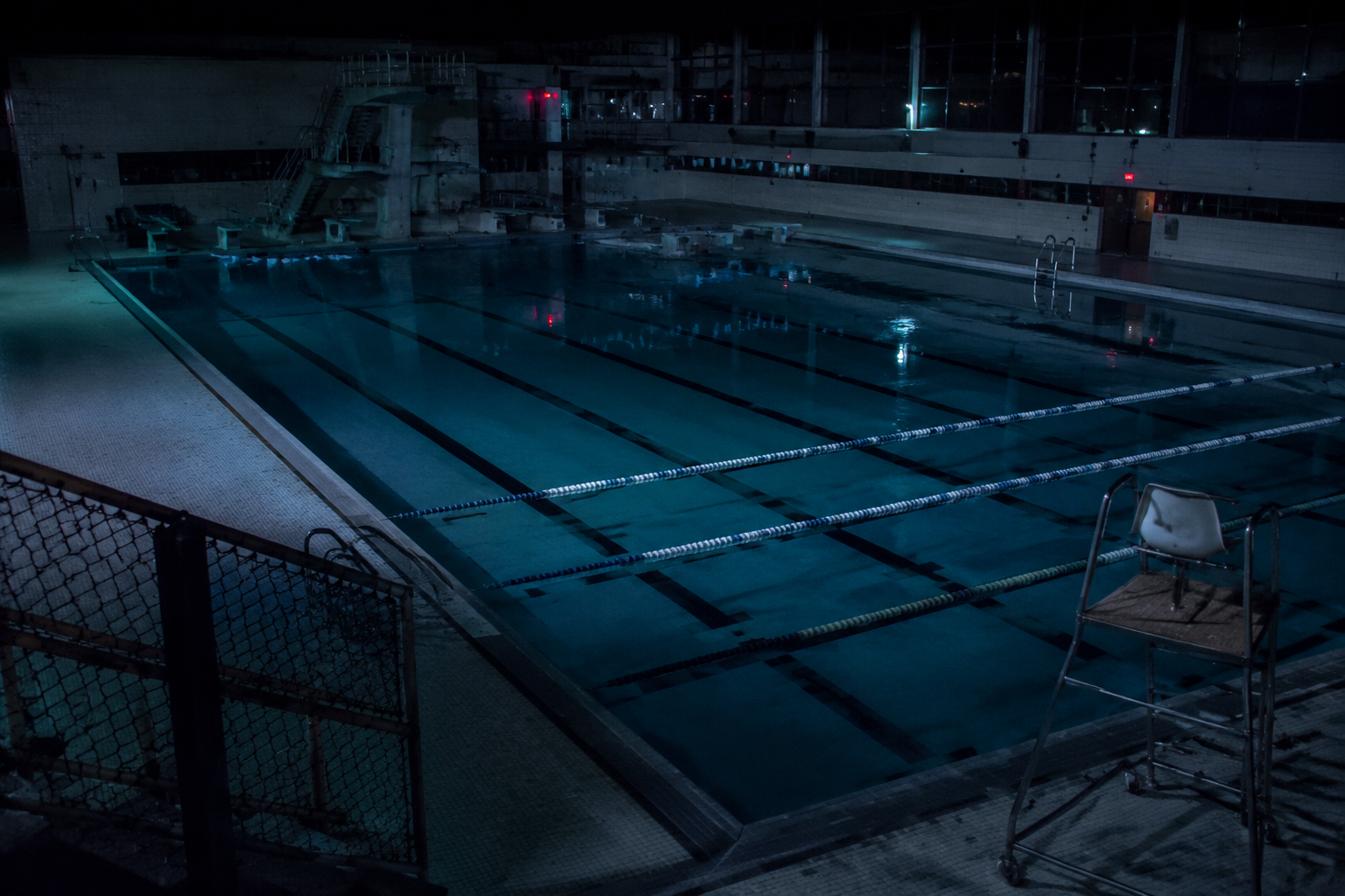 Empty municipal natatorium at night with lane ropes across a still lap pool and a lifeguard chair in dim light.