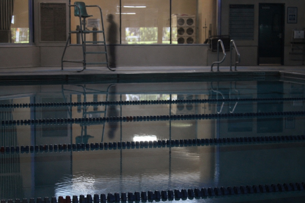 Still pool water reflects a shadowy figure near a lifeguard chair in an otherwise empty natatorium.