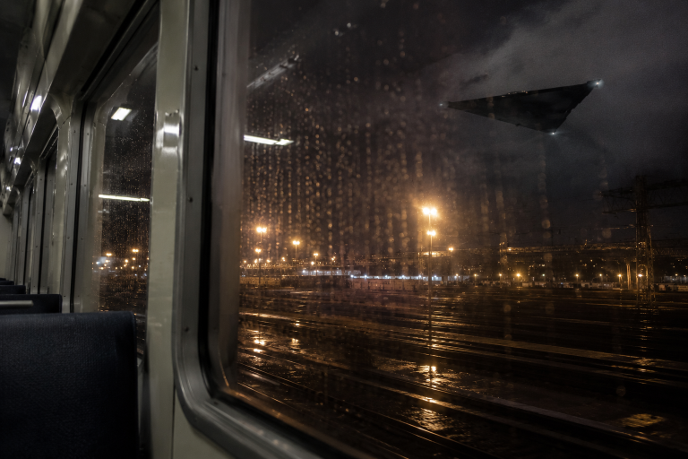 Rain streaks cover a commuter train window while a black triangular object hangs over a rail yard.