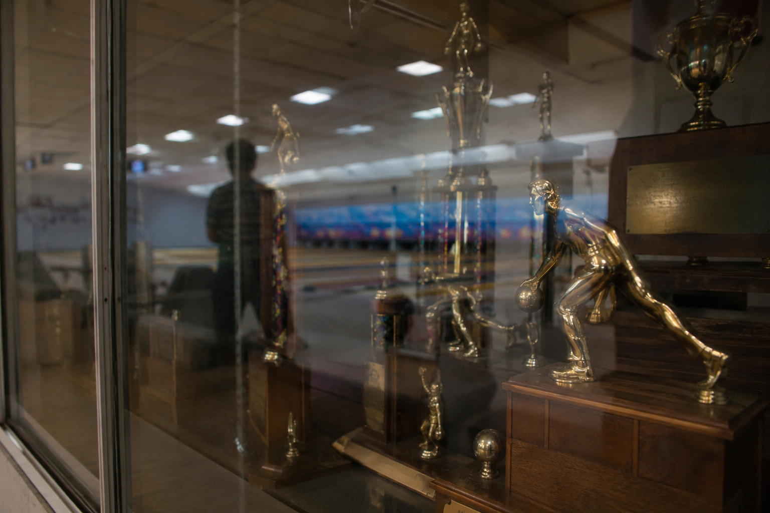 Trophy case reflection with a faint figure near closed bowling lane doors