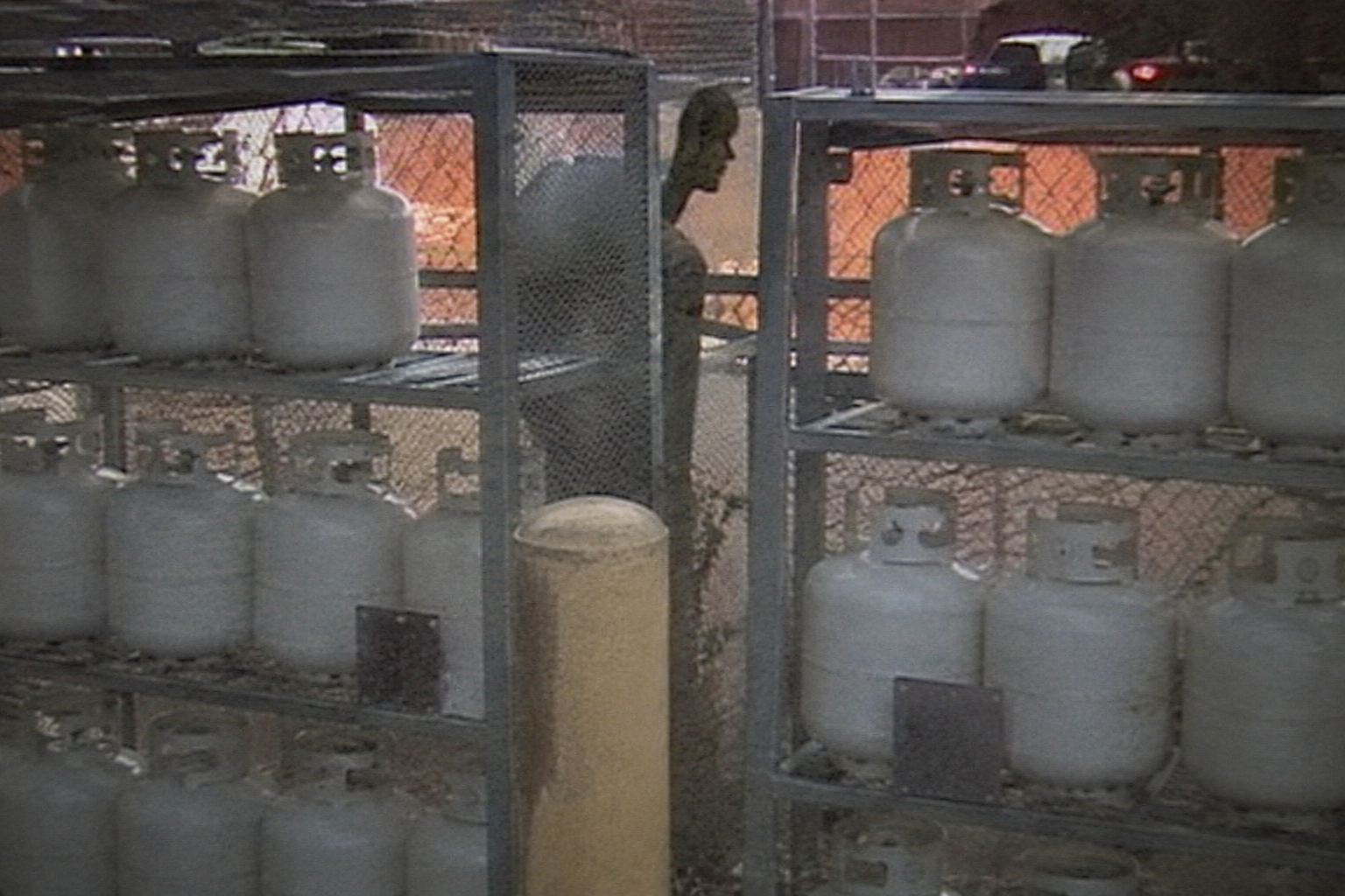 Thin unknown visitor partly hidden behind propane tank racks in security footage.